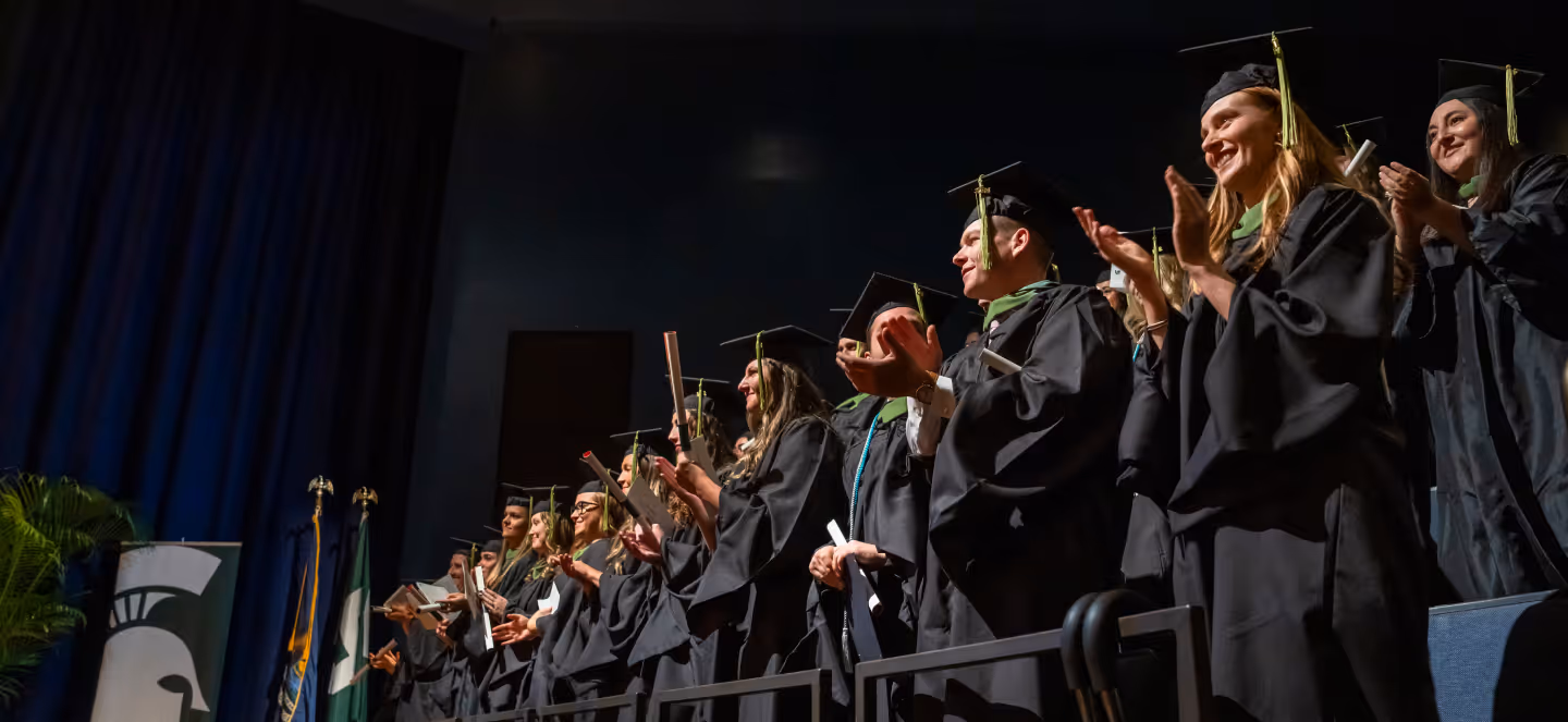 Graduates in black caps and gowns stand on stage applauding during a commencement ceremony, holding diplomas as Michigan State University banners and flags appear in the background.