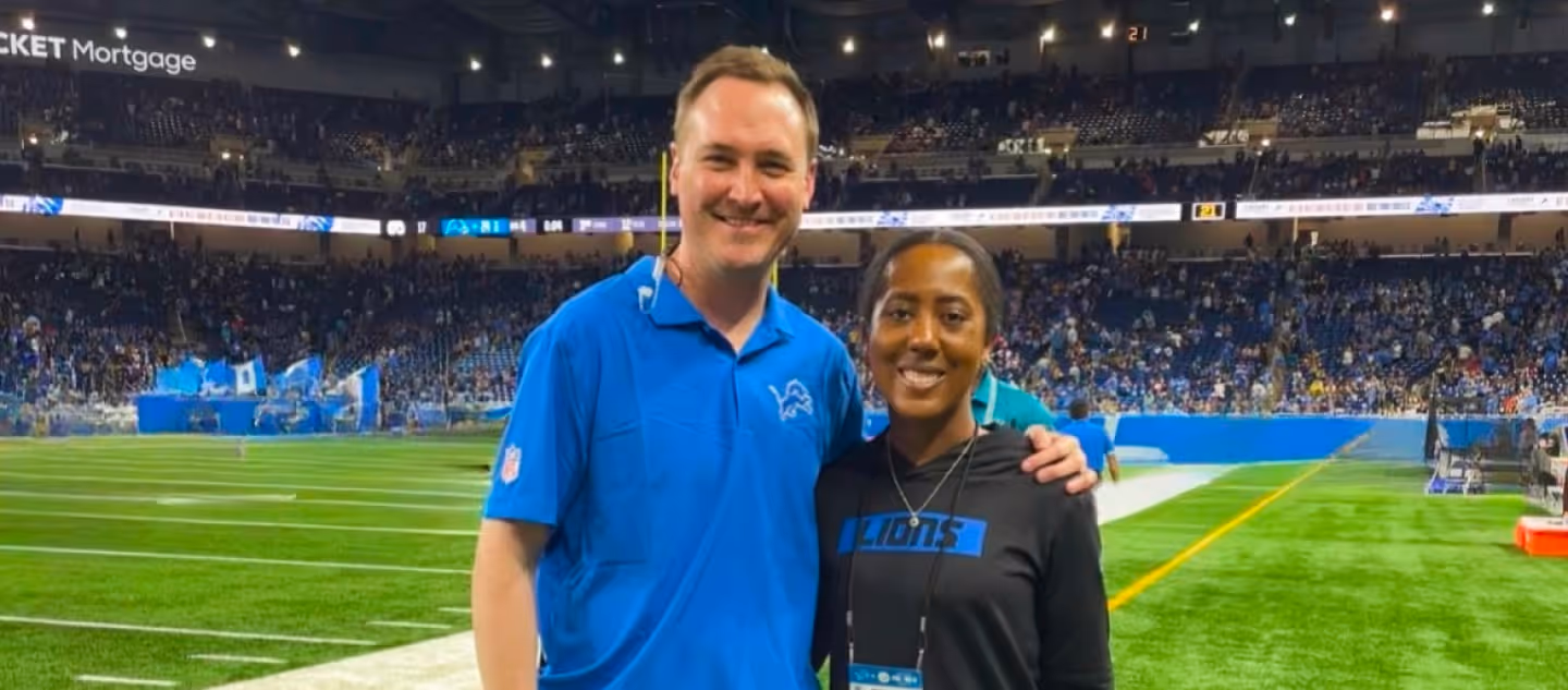 Two people smiling on a football field inside a packed indoor stadium, with one wearing a blue team polo and the other in a black hoodie, standing near the sideline before or during a game.