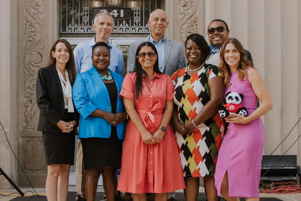 Group of community and organizational leaders standing together outside a historic building entrance, smiling for a group photo during a public event or announcement.