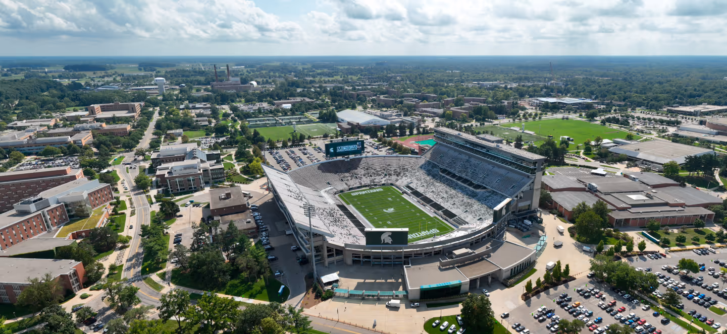 Aerial view of Michigan State University’s Spartan Stadium and surrounding campus, showing the football field, seating bowl, nearby academic buildings, parking areas, and green spaces under a partly cloudy sky.