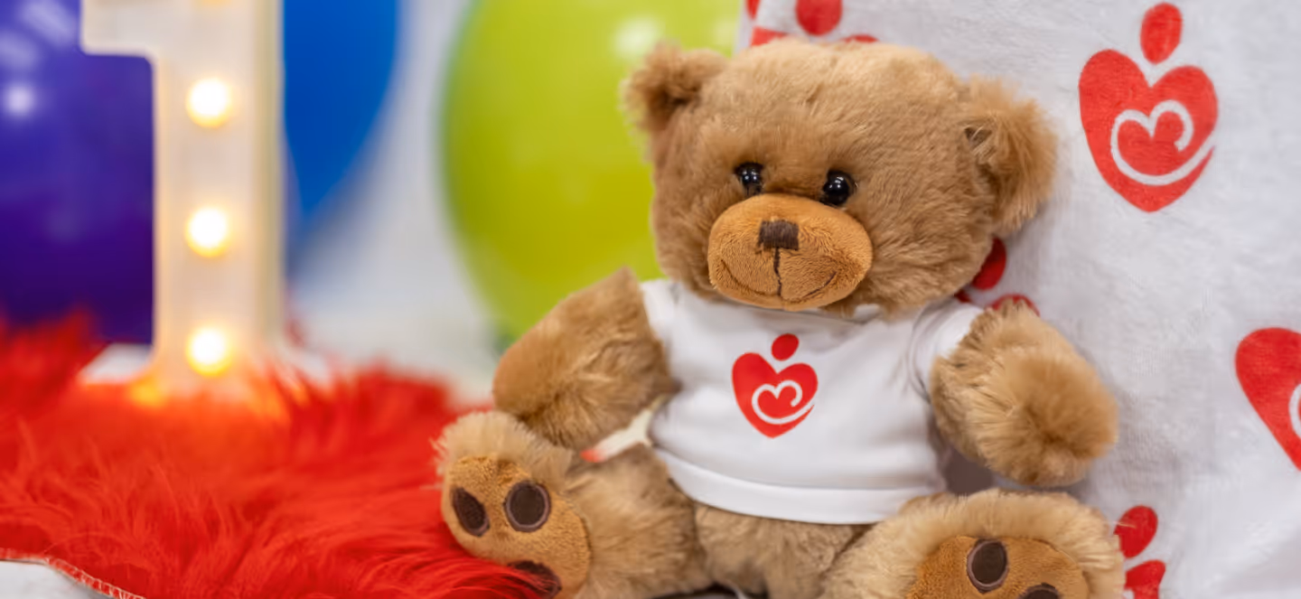 Plush teddy bear wearing a white shirt with a red heart logo, seated among colorful balloons and soft decorations.