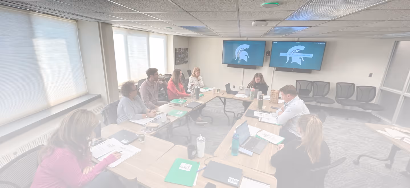 Group of staff seated around tables in a conference room, participating in a meeting with laptops, notebooks, and screens displaying the Michigan State University Spartan logo.