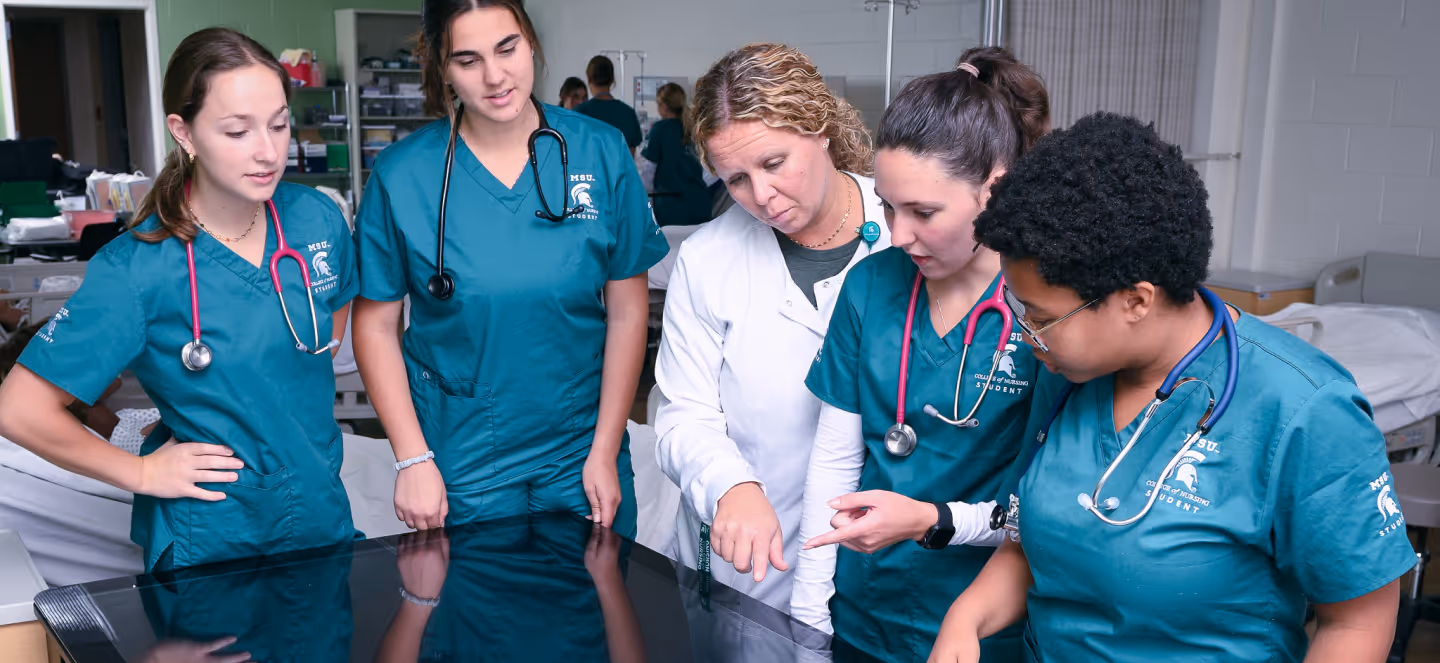 Group of nursing students in clinical scrubs gathered around an instructor, reviewing information on a large touchscreen in a clinical skills lab.