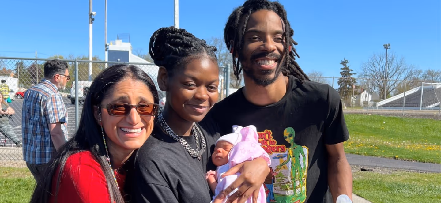 Three adults smiling outdoors, one holding a baby, with buildings and a grassy area visible in the background.