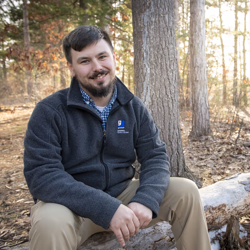 Bailey LaPan, a homeless services manager, sitting on a log in the woods facing the camera.