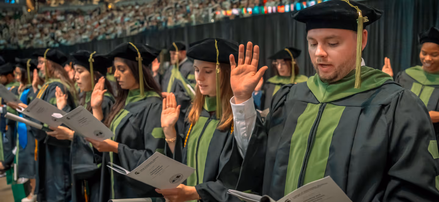 Graduates in caps and gowns seated at a commencement ceremony, raising their right hands during an oath or pledge.