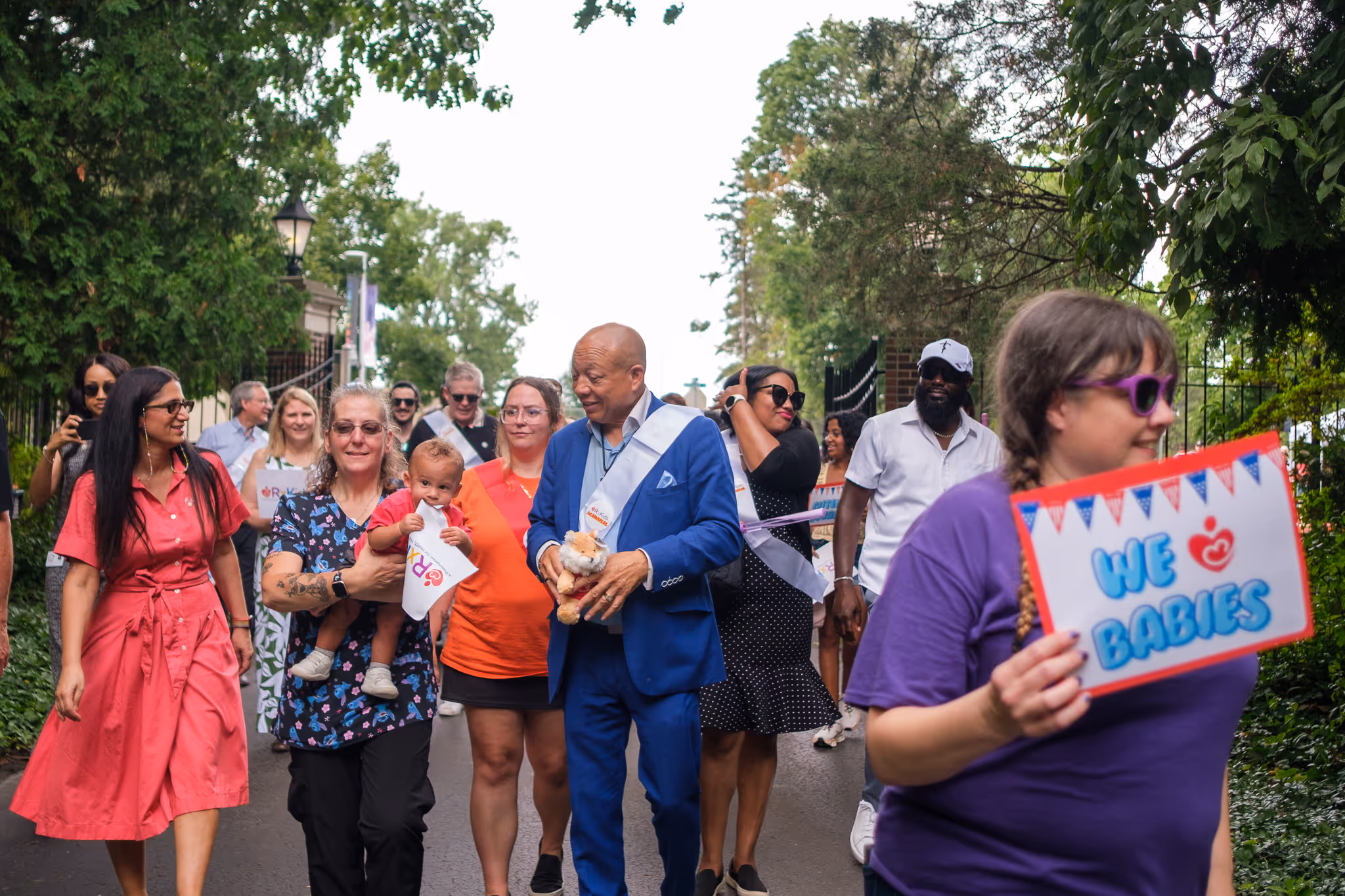 Group of adults and children gathered outdoors at a community event, one person holding a colorful handmade sign, smiling and engaging with the crowd.