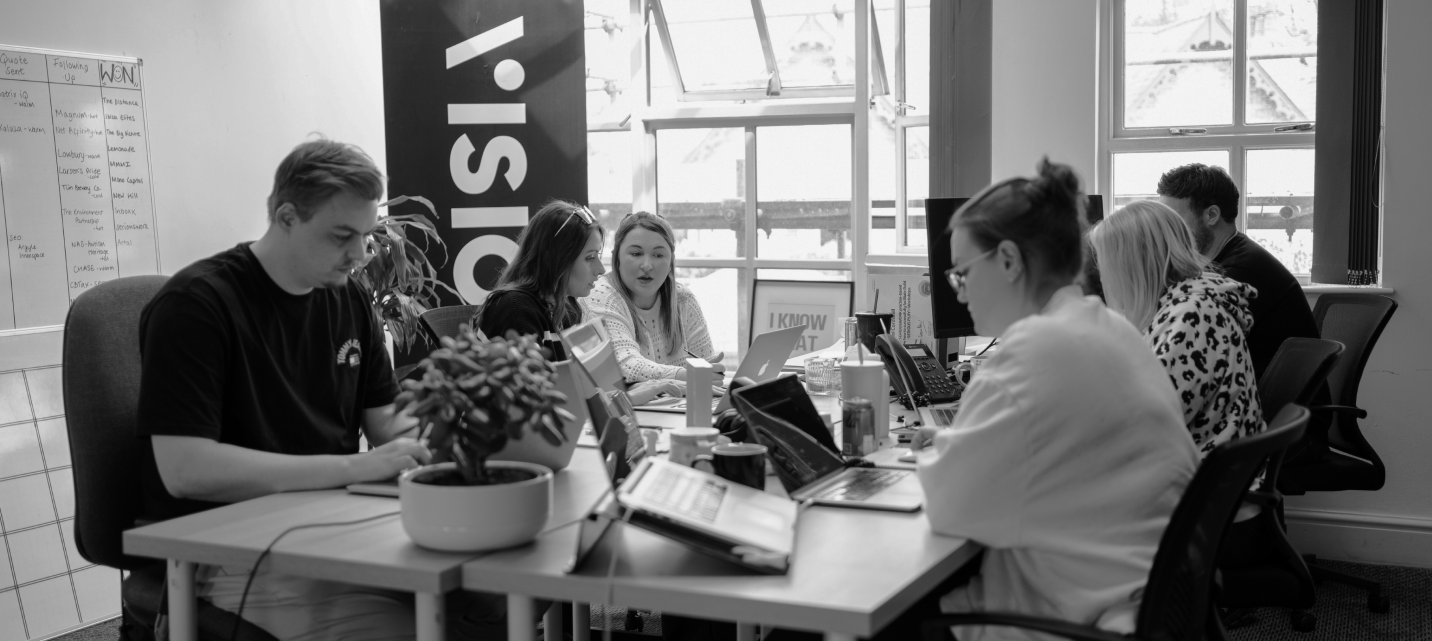 Six people collaborating around a table with laptops and notebooks in a bright office space.