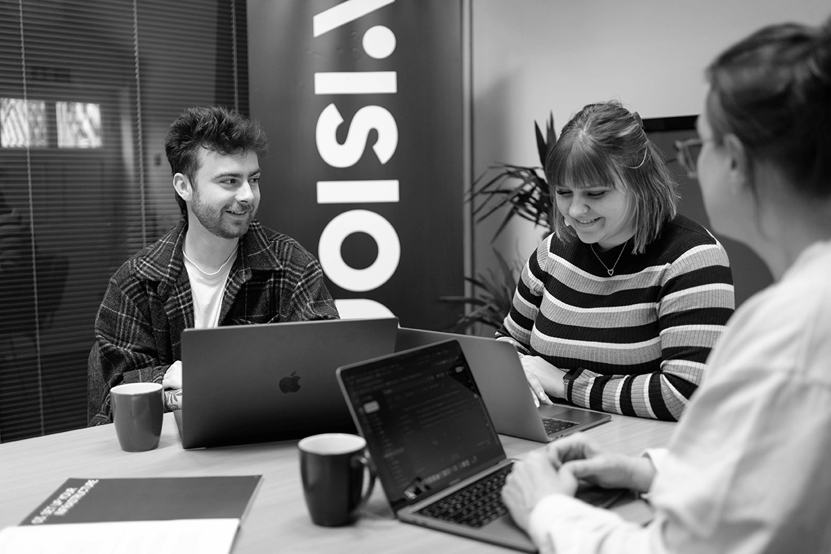 Three people sitting around a table working on laptops and smiling in a modern office meeting room.