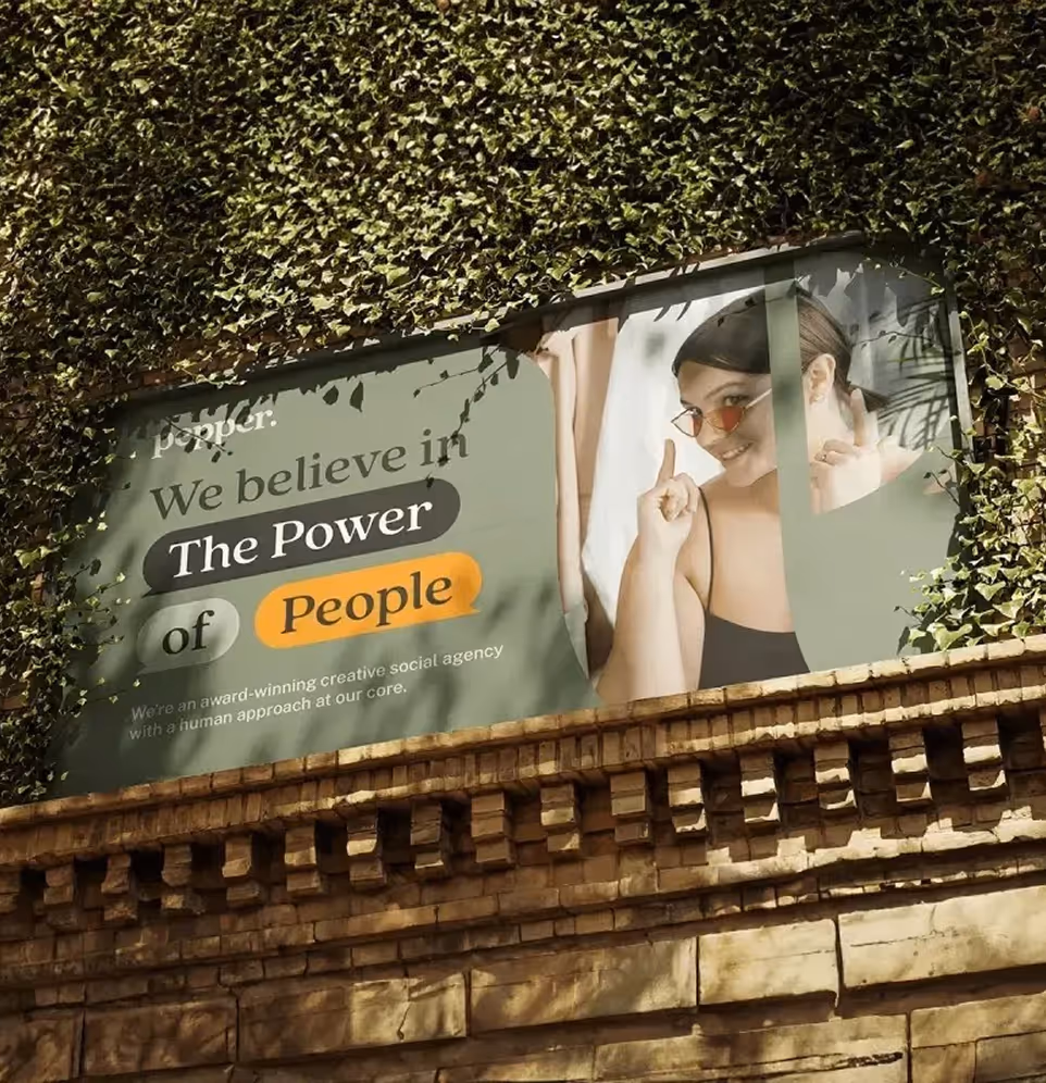 Outdoor billboard on a brick wall with ivy, showing a woman smiling and pointing upward next to text that reads, 'We believe in The Power of People.'