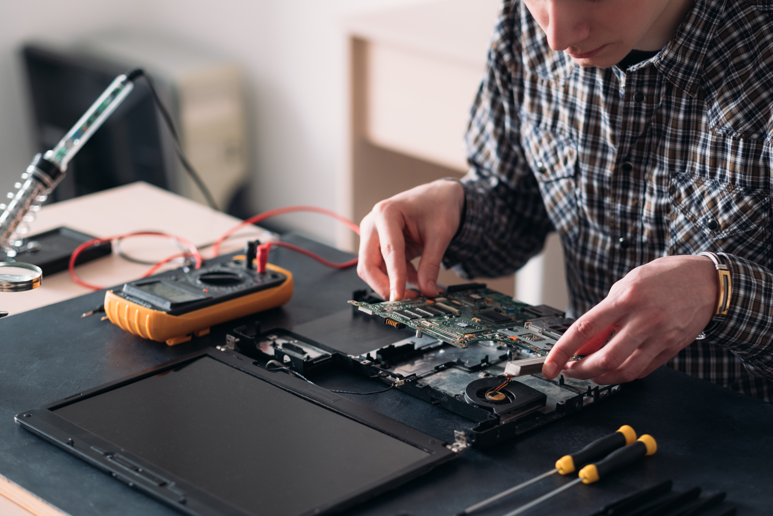 A man in a flannel shirt repairing a laptop on a table, as part of the refurbishing stage by laptop recycling services in Singapore.