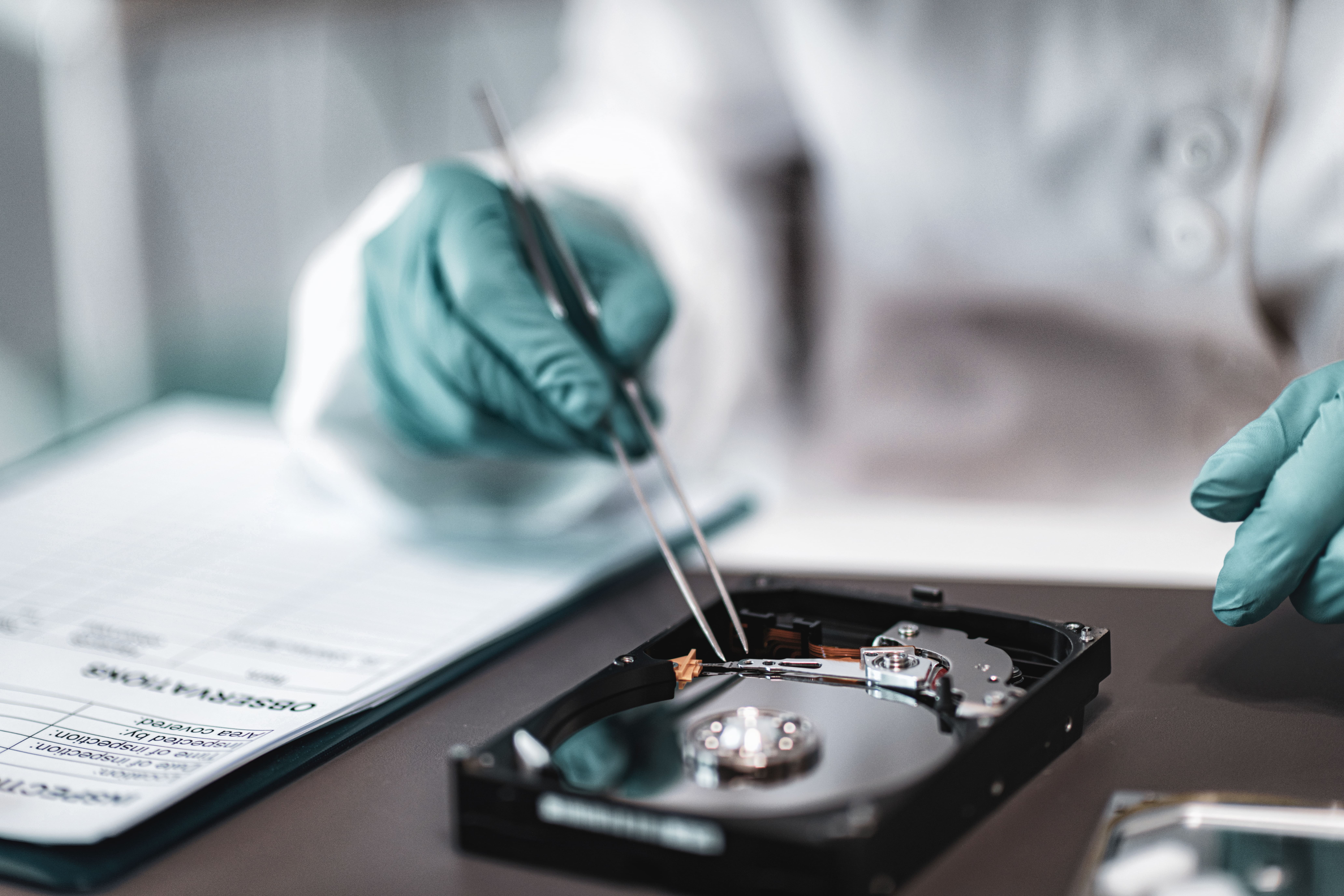 Gloved forensic examiner inspects an opened hard drive with tweezers beside an evidence form