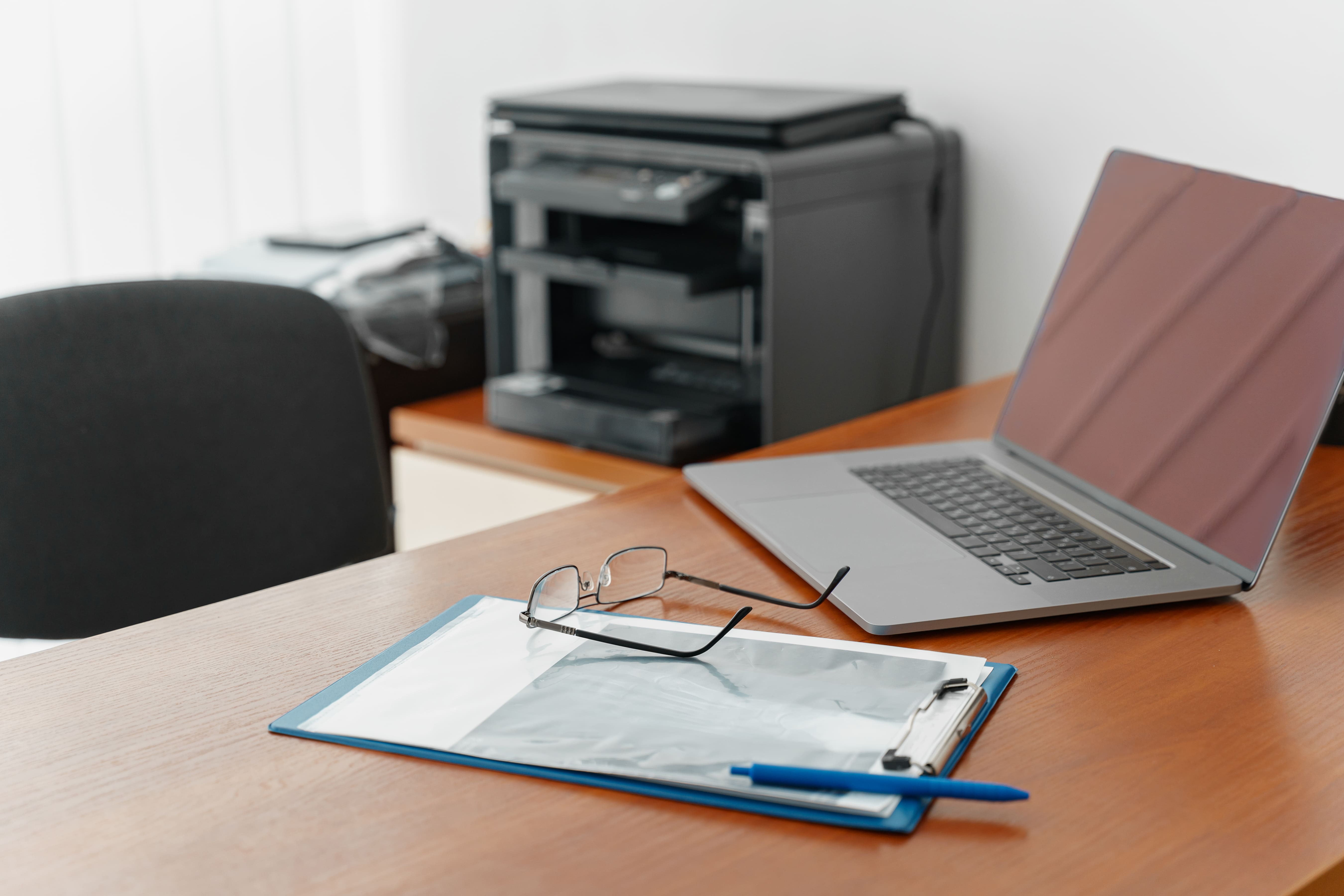 Blue clipboard with glasses and pen beside a laptop on an office desk, with a printer in the background