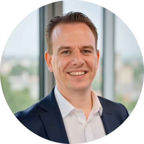 Smiling man wearing a white shirt and dark blazer standing in front of a blurred window background.