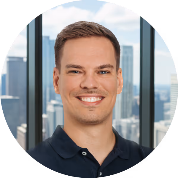 Smiling man with short brown hair wearing a dark polo shirt, with a cityscape visible through large windows behind him.