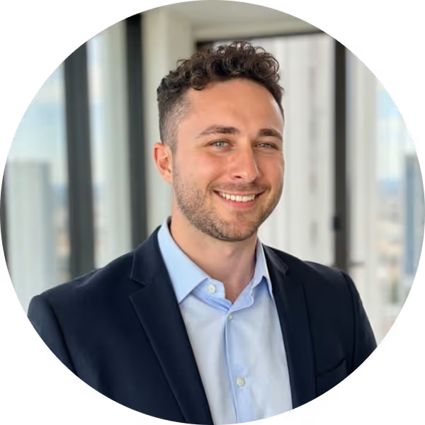 Smiling man with curly hair and beard wearing a dark blazer and light blue shirt in an office with large windows.