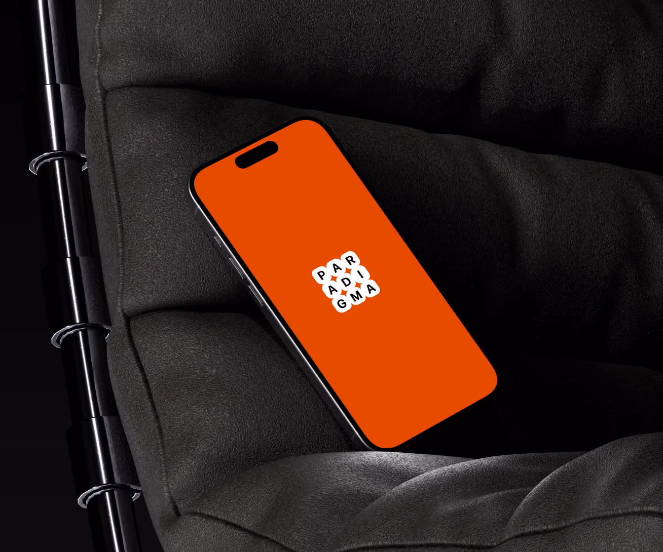 Smartphone with a red screen displaying the word 'PARADIGMA' in white bubble letters placed diagonally, resting on a black cushioned chair with a metal frame.