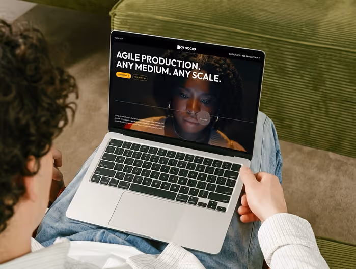 Person holding a laptop open on their lap, showing a website with the text 'Agile production. Any medium. Any scale.' and a reflected face of a woman on the screen. The person is sitting on a green couch wearing a light-colored shirt and jeans.