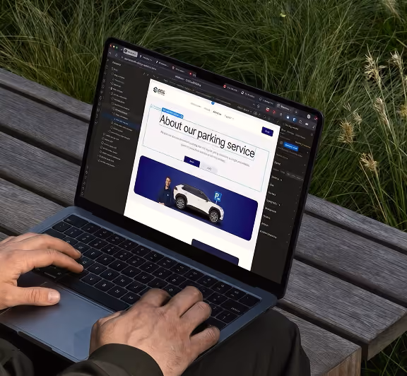 Person typing on a laptop keyboard while sitting on a wooden bench next to tall green grass, with the laptop screen displaying a webpage about a parking service featuring a white car image and text.