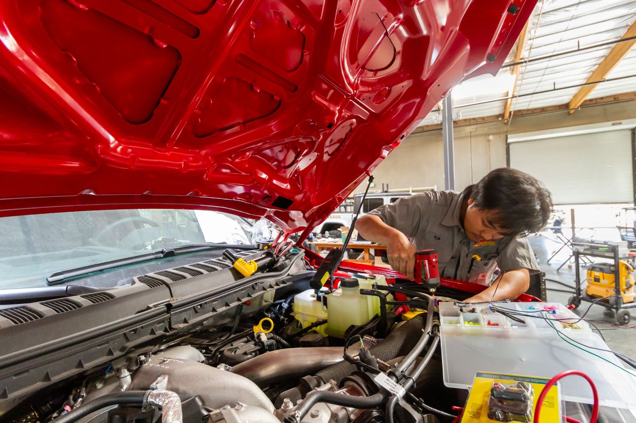 Mechanic working under hood of red truck inside repair shop, using power tool and wiring kit for engine diagnostics and maintenance.