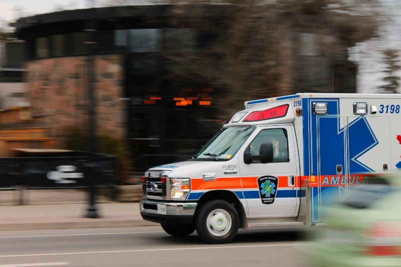 An ambulance from Paramedic Services speeds down a city street with emergency lights on, passing blurred buildings and traffic.