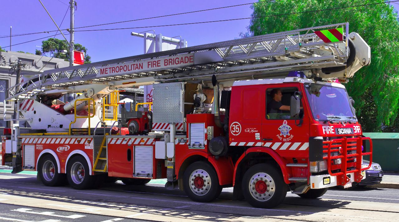 Red Metropolitan Fire Brigade ladder truck parked roadside, featuring extended aerial ladder, emergency rescue equipment, and multiple heavy-duty axles.