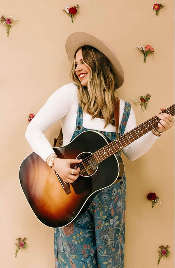 A young white woman with long blonde hair holds a guitar.
