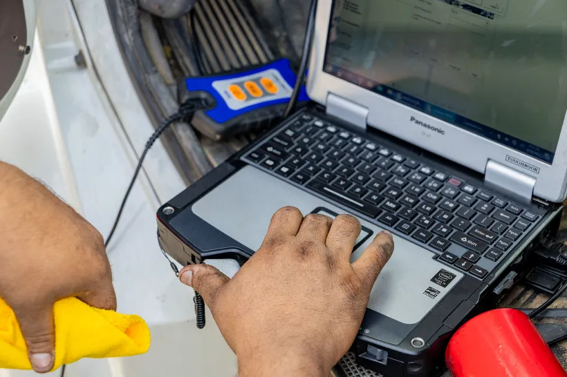 Technician diagnosing electrical problems using a rugged Panasonic laptop connected to vehicle diagnostic tools.