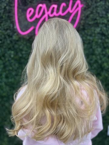 Person with long, wavy blonde hair standing with back to the camera in front of a green leafy wall with pink neon sign reading 'Legacy'.