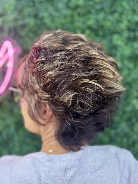 Back view of a person with short, curly, highlighted hair styled in a textured updo wearing glasses and a light gray top.