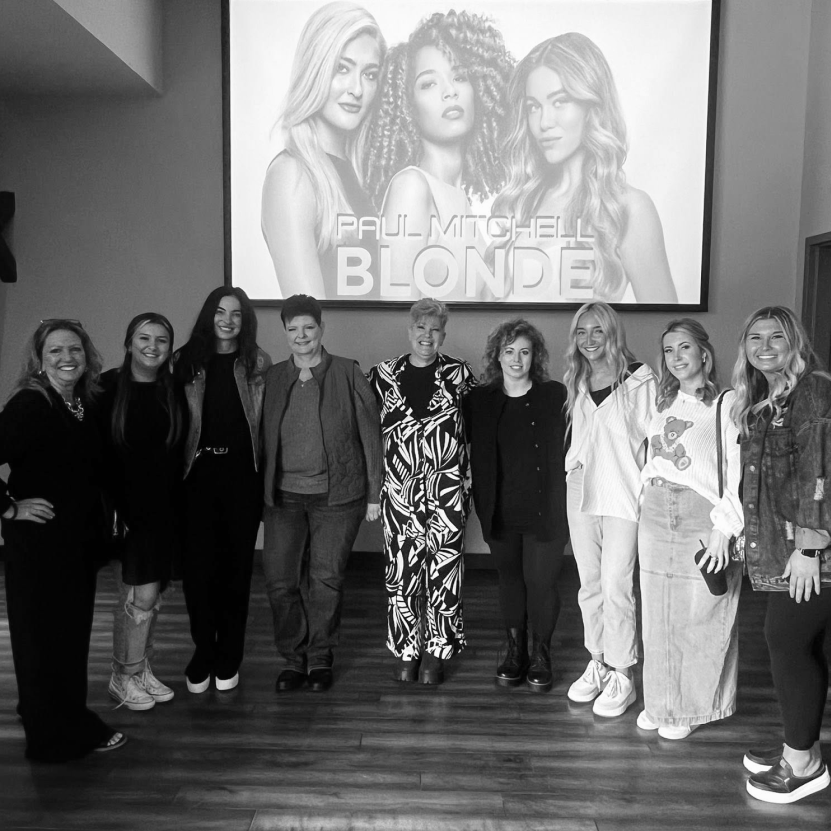 Group of ten women standing side by side smiling in front of a large poster that says 'Paul Mitchell Blonde' featuring three blonde models.