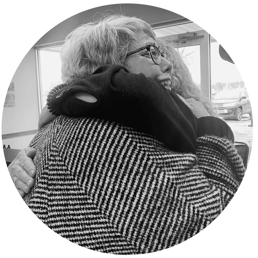 Two women warmly hugging indoors near a window, one wearing glasses and a houndstooth patterned coat.