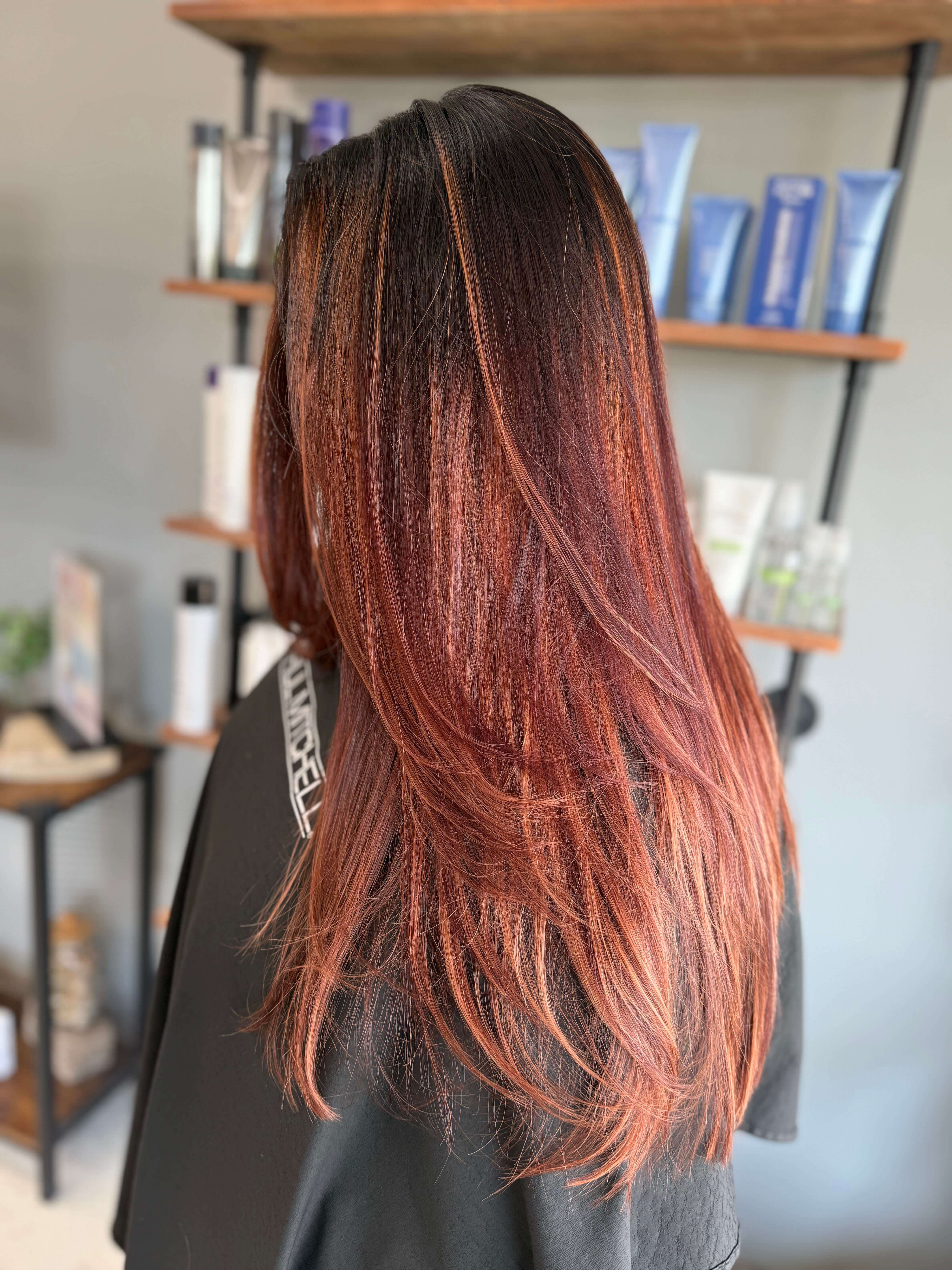 Person with long, layered hair dyed in dark brown and reddish hues, standing in a salon with hair products on shelves in the background.