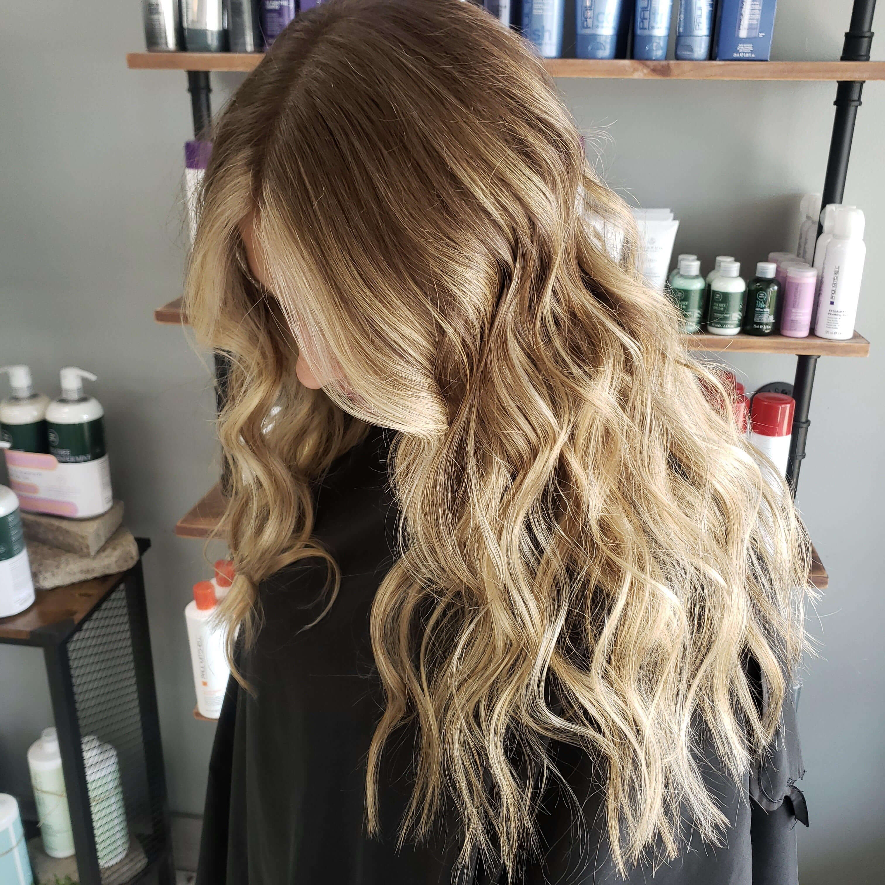 Person with long, wavy, blonde balayage hair styled in loose curls in a salon setting.