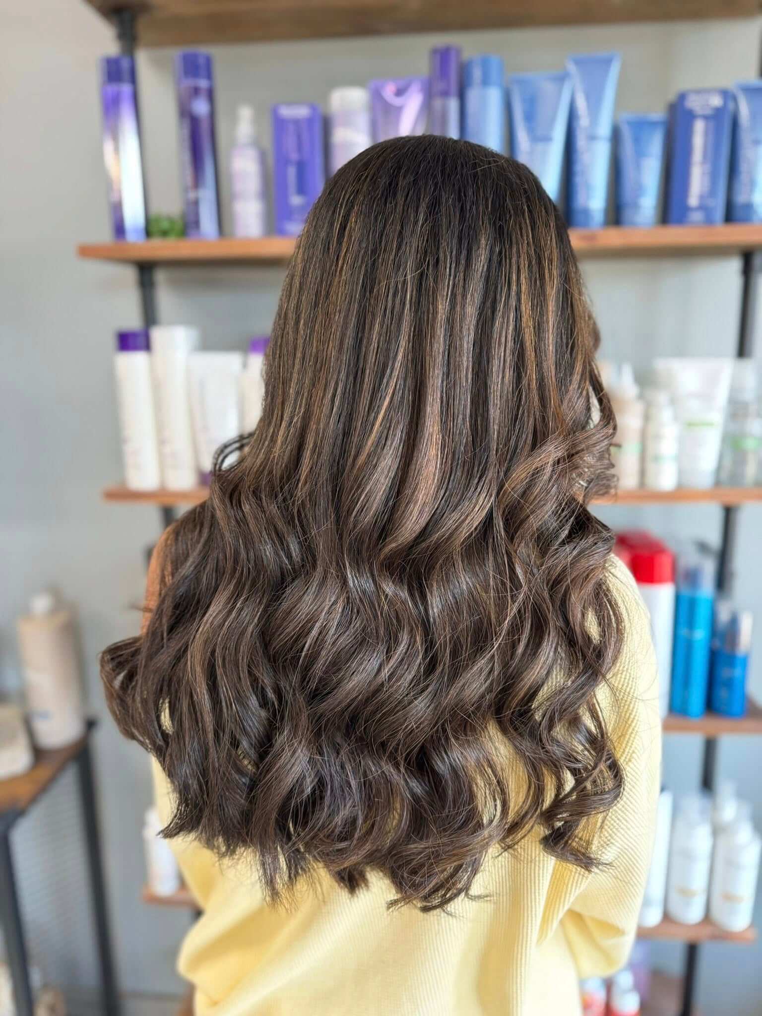 Back view of a person with long, thick, wavy dark brown hair with subtle highlights standing in front of shelves with hair care products.