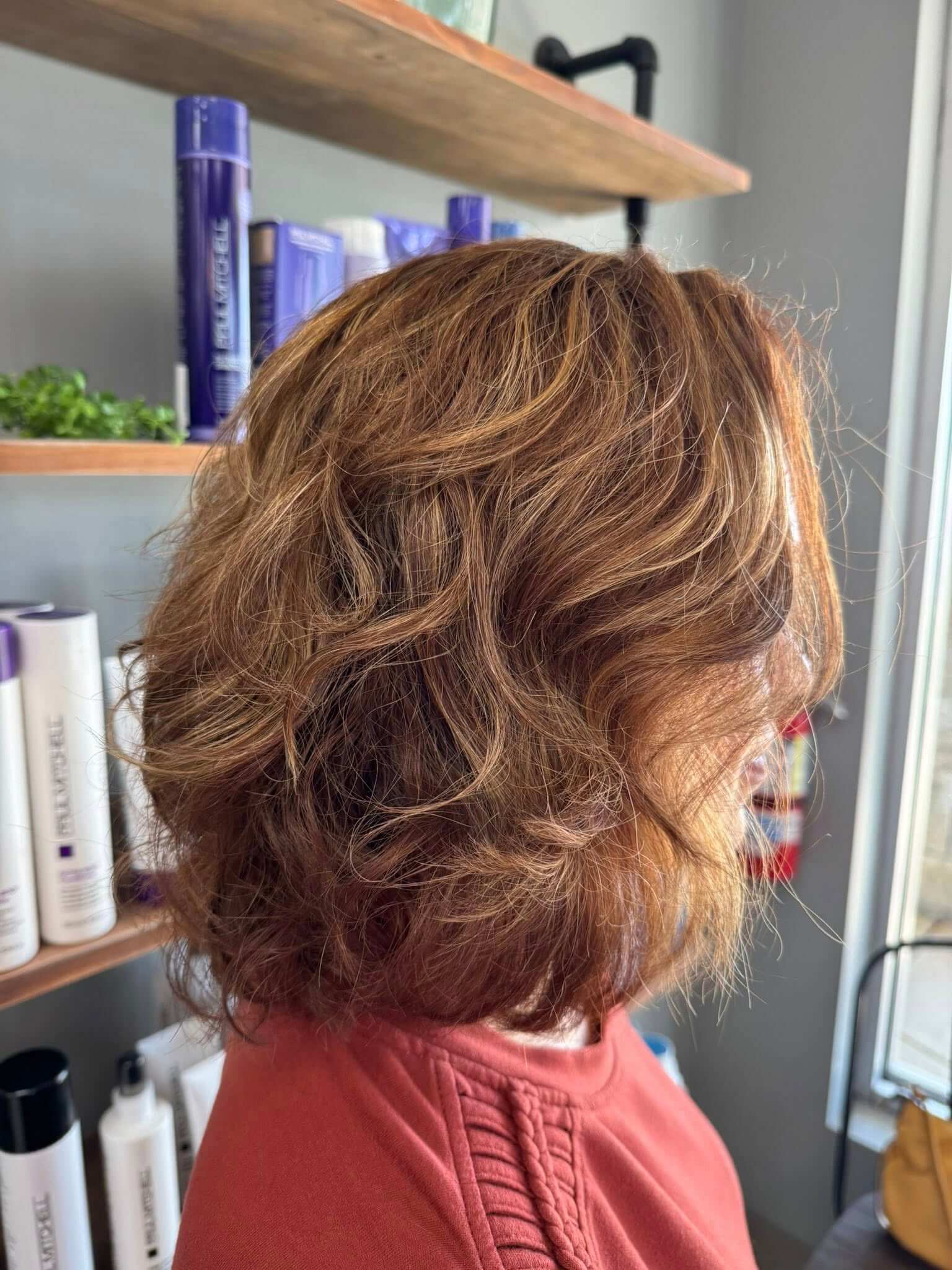 Side view of a person with medium-length, wavy light brown hair and subtle highlights, wearing a rust-colored top in a salon setting.