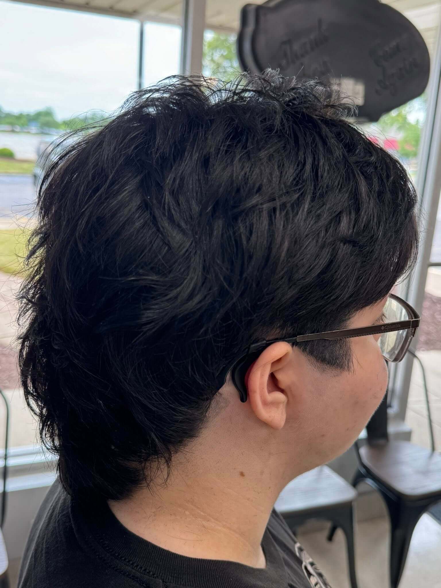 Side profile of a person with short, thick black hair wearing glasses and a black shirt inside a building.