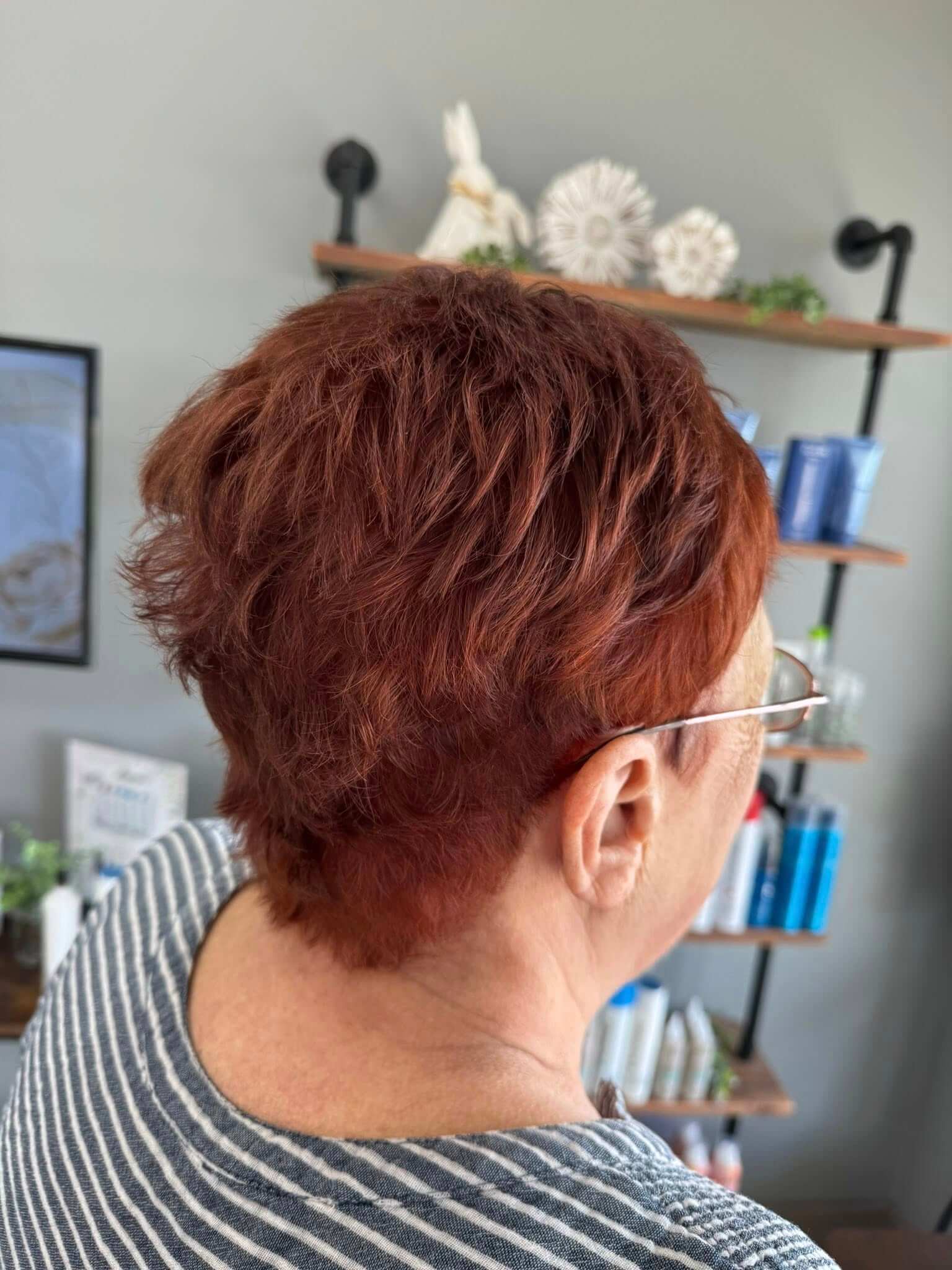 Back view of a person with short, curly, reddish-brown hair wearing glasses and a striped gray top in a salon setting.
