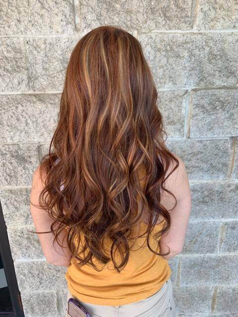 Woman with long, wavy, brown hair with blonde highlights standing against a gray stone wall.
