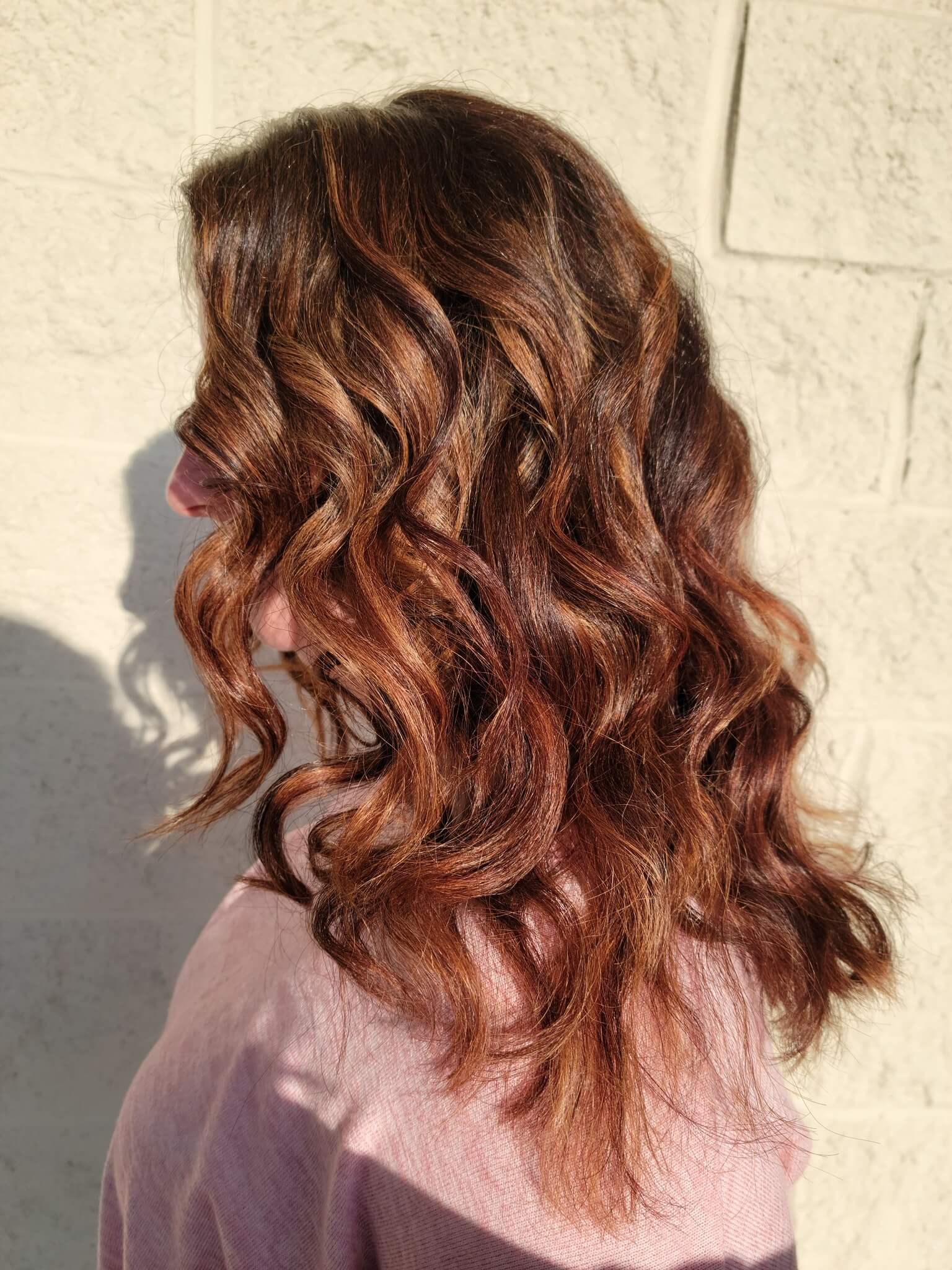 Side profile of a person with shoulder-length, voluminous chestnut brown hair styled in loose waves, wearing a light pink top.