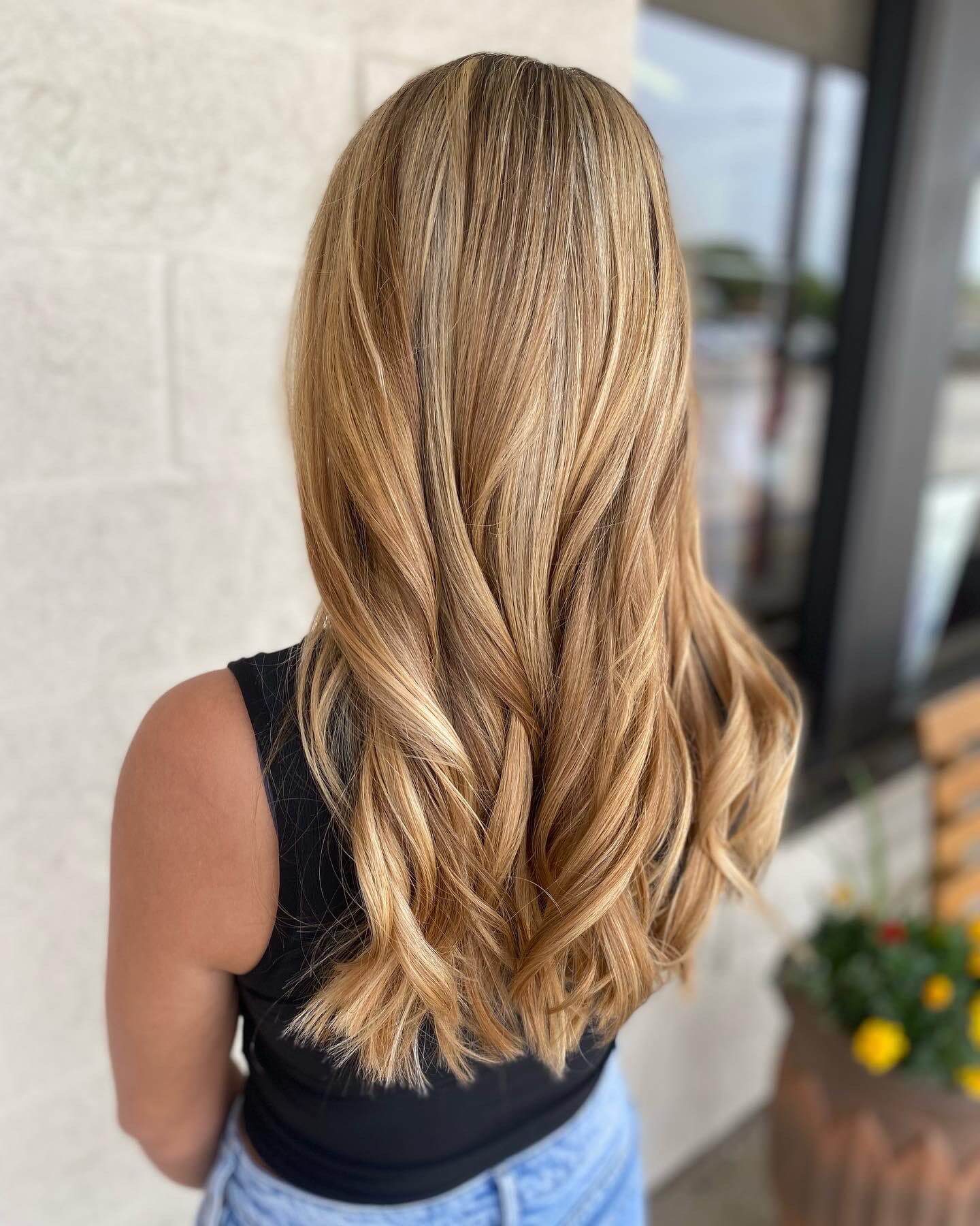 Back view of a person with long, wavy blonde hair wearing a sleeveless black top and light blue jeans.