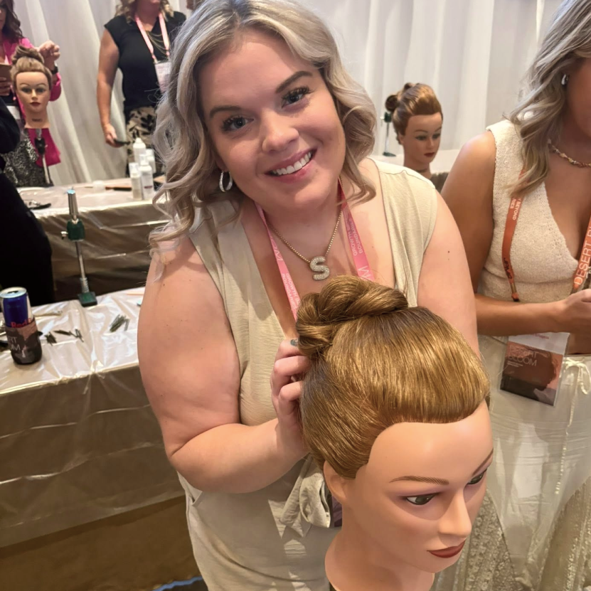 Woman smiling while styling hair on a mannequin head with an updo hairstyle at a hair education event.