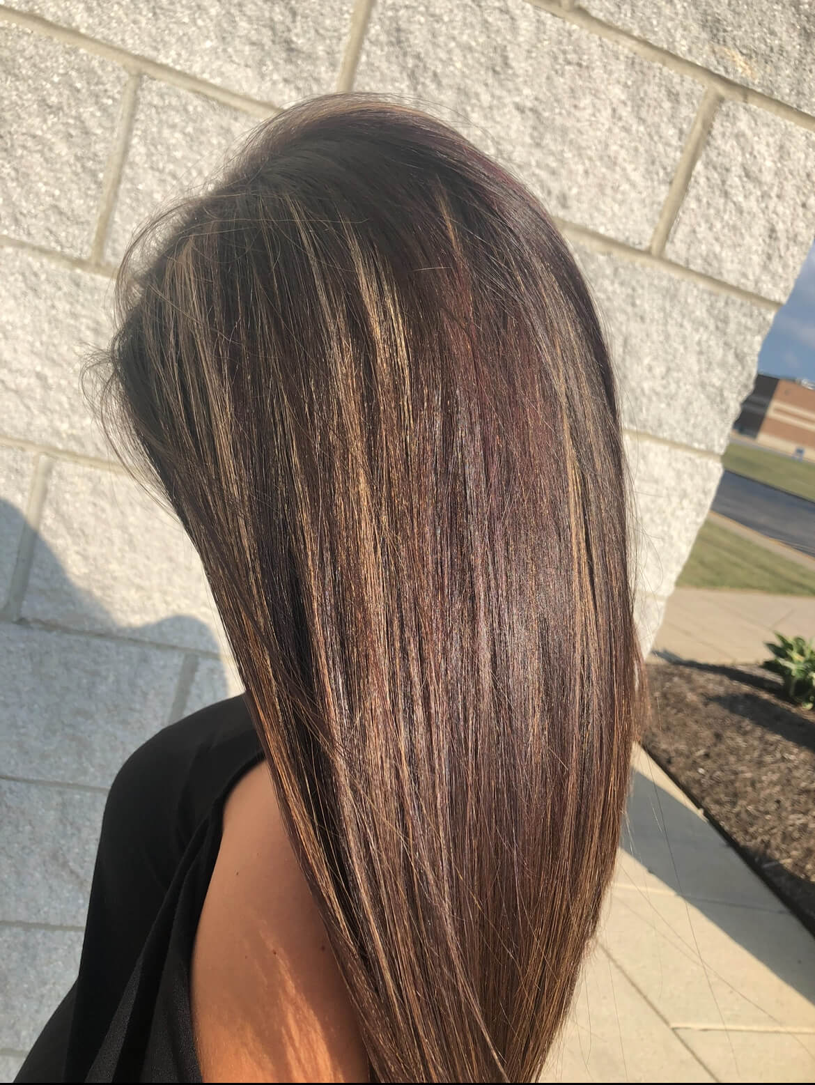 Woman with long, straight brown hair featuring subtle blonde and light brown highlights, photographed outdoors near a light brick wall.