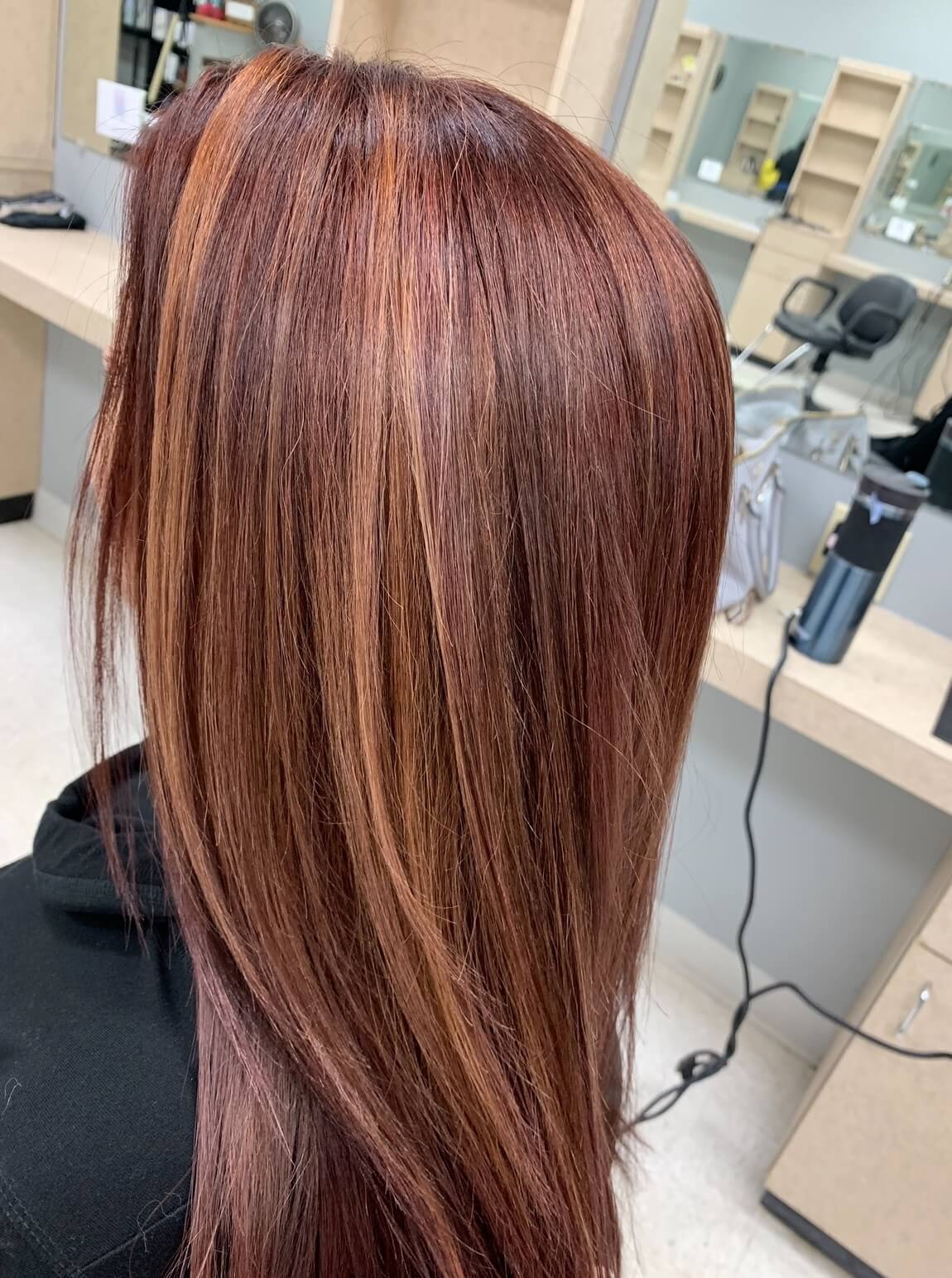 Person with straight, shoulder-length hair dyed in warm reddish-brown tones with lighter highlights, viewed from the back in a salon setting.