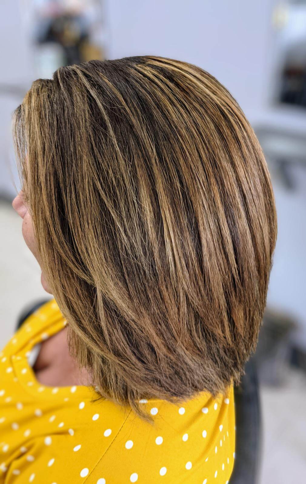Close-up side view of a woman with a layered bob haircut featuring light brown and blonde highlights, wearing a yellow polka dot top.