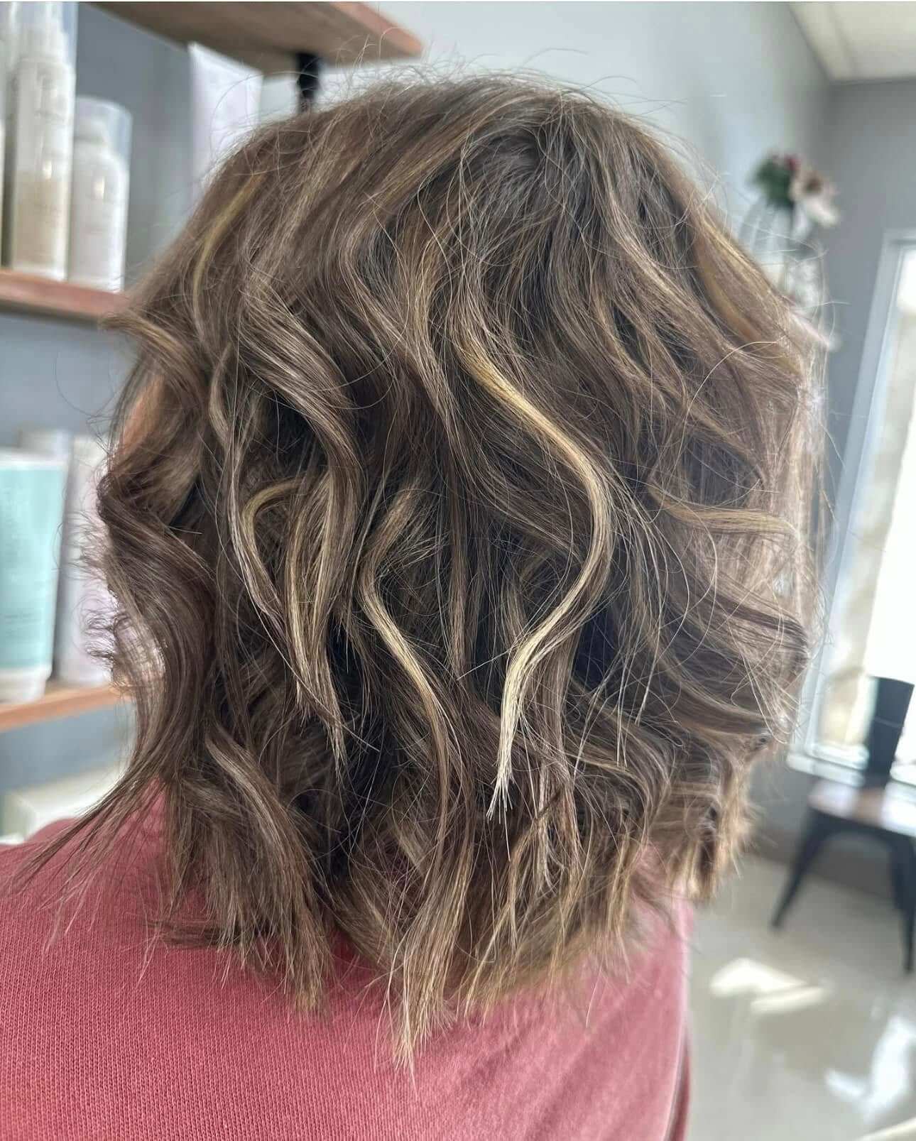 Back view of medium-length wavy brown hair with lighter highlights, indoors with shelves and a window in the background.