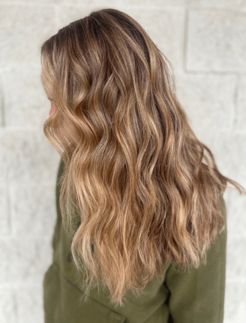 Person with long, wavy, highlighted brown and blonde hair wearing a green jacket, facing away from the camera.