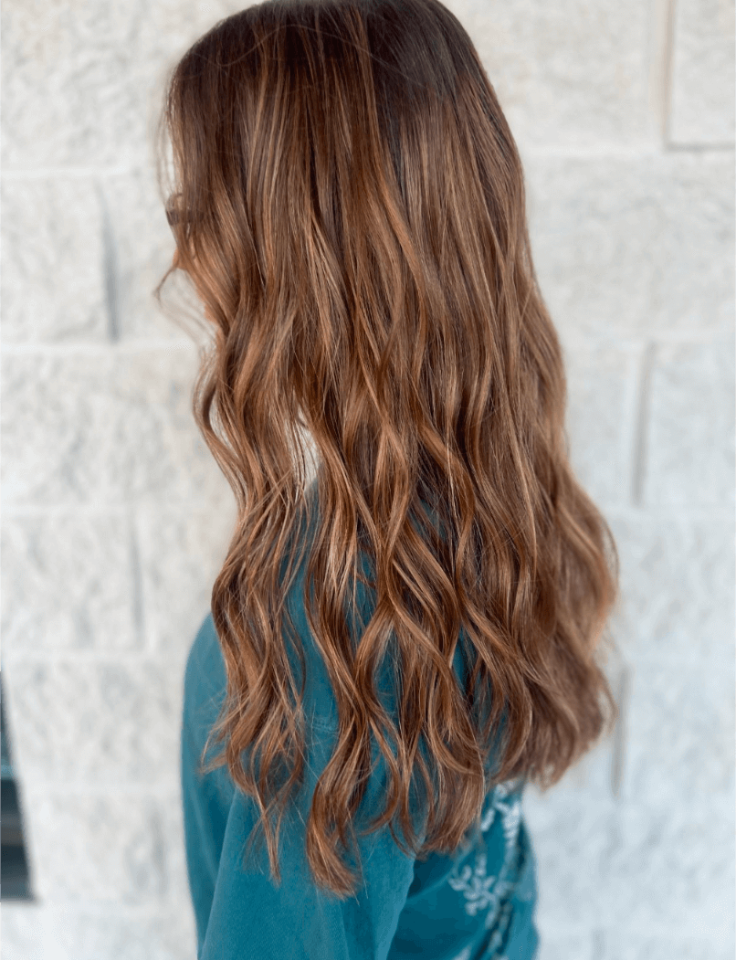 Side view of a person with long, wavy brown hair wearing a teal top standing in front of a light gray stone wall.