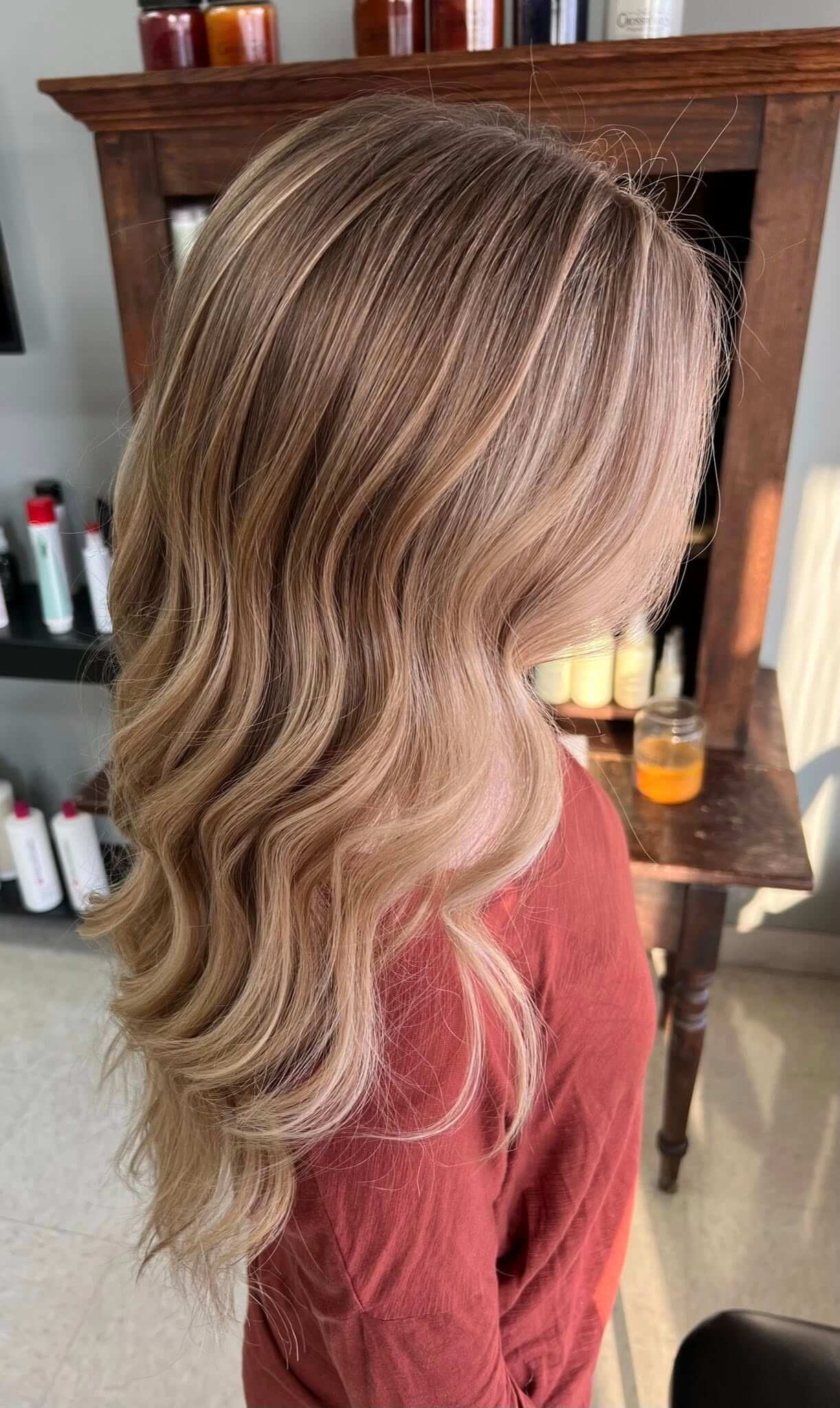 Side view of long, wavy blonde hair with highlights on a person wearing a rust-colored top.