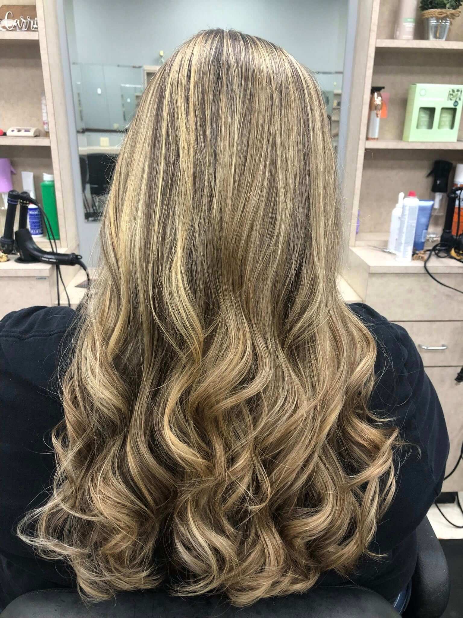 Back view of long, wavy blonde hair with darker roots in a salon setting.